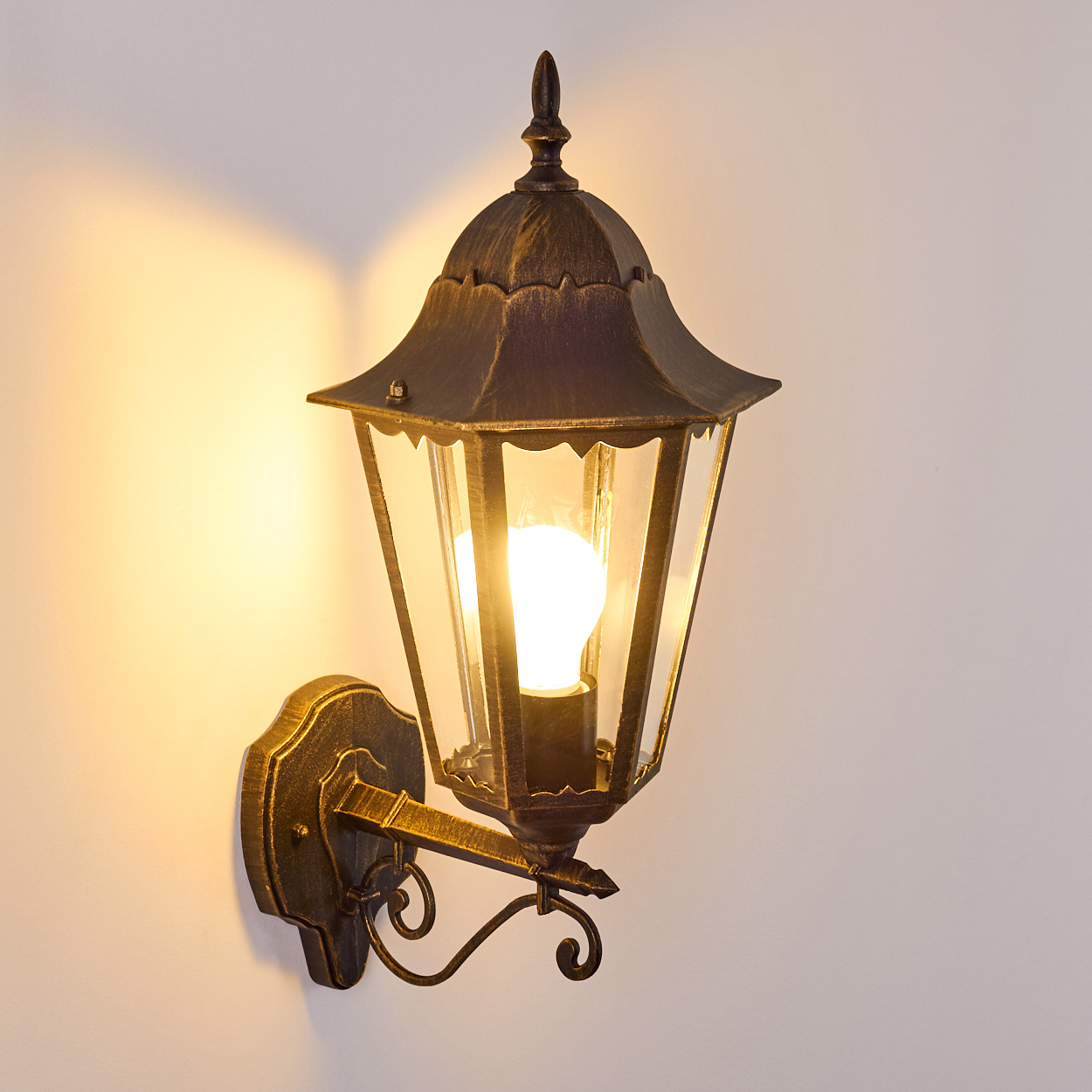 Wandlampe mit goldfarbenem Rahmen, Glasscheiben und Glühbirne. An einer weißen Wand.
