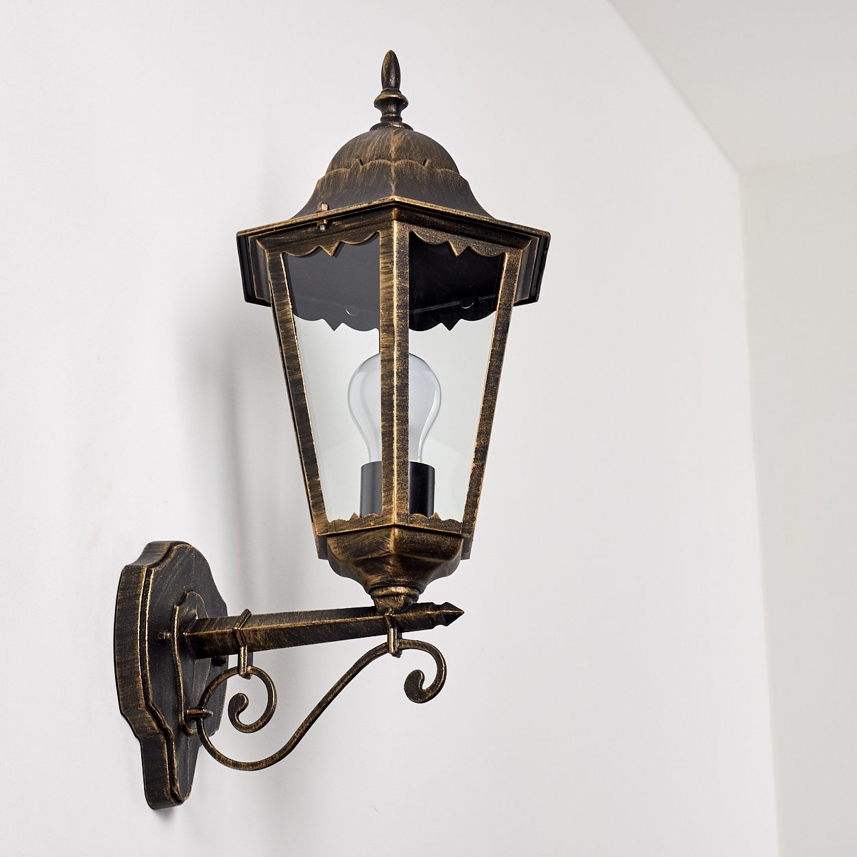 Wandlampe mit goldenem Rahmen, Glasscheiben und Glühbirne. An einer weißen Wand.