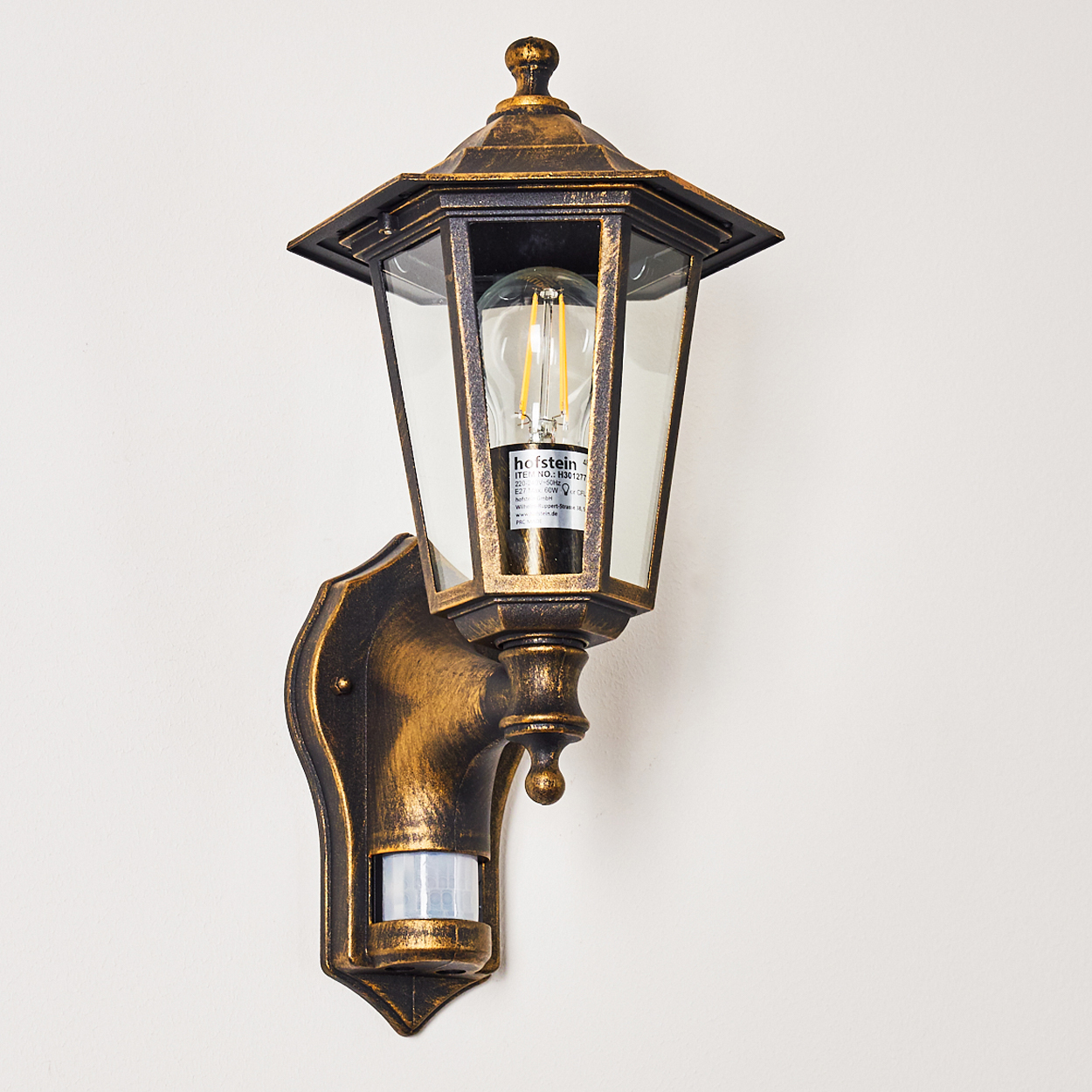 Wandmontierte, verzierte Außenlampe, beleuchtet mit sichtbarer Glühbirne, auf neutralem Hintergrund.