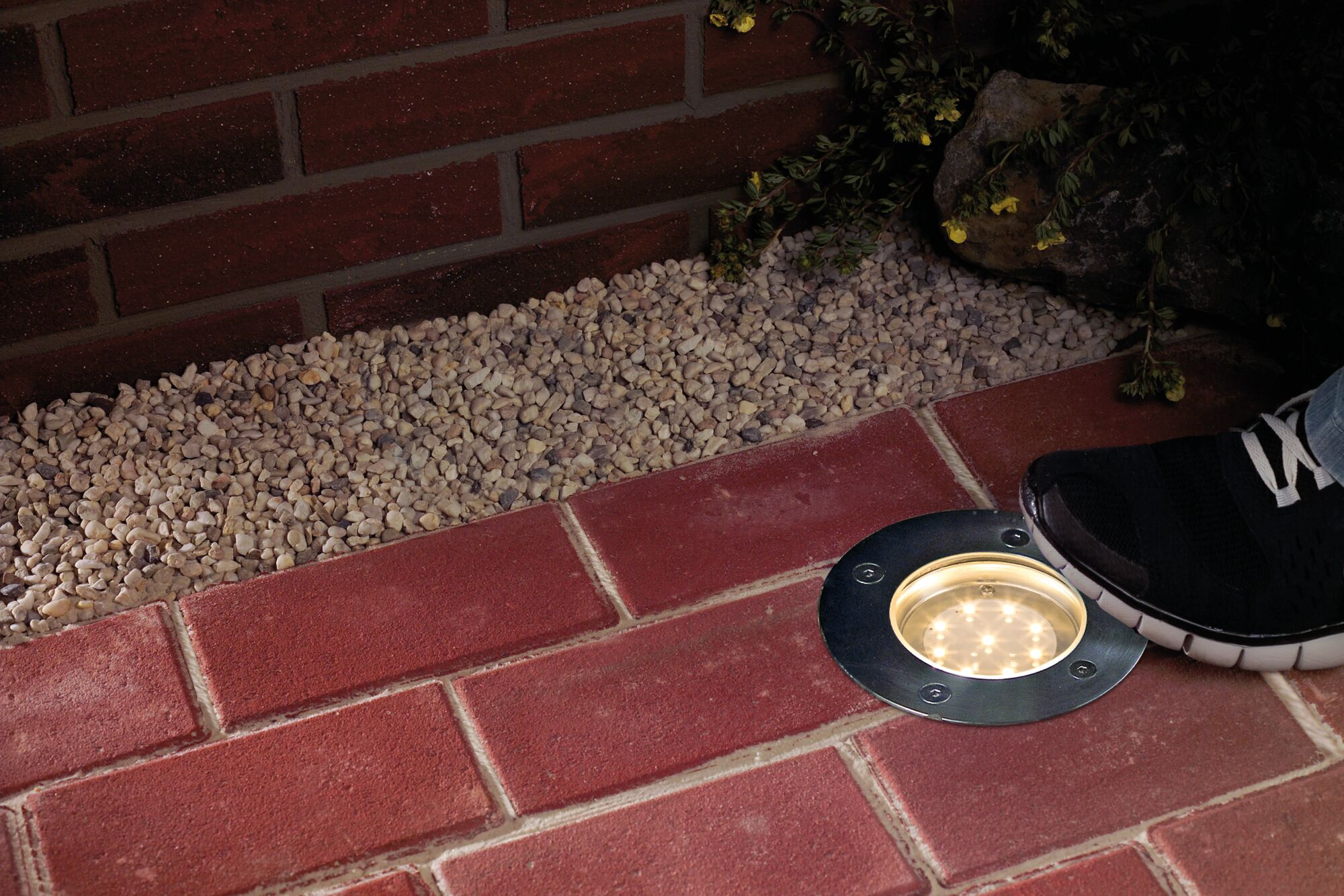 Un entorno al aire libre. Un patio de ladrillos, grava y una luz LED empotrada con un pie cerca.