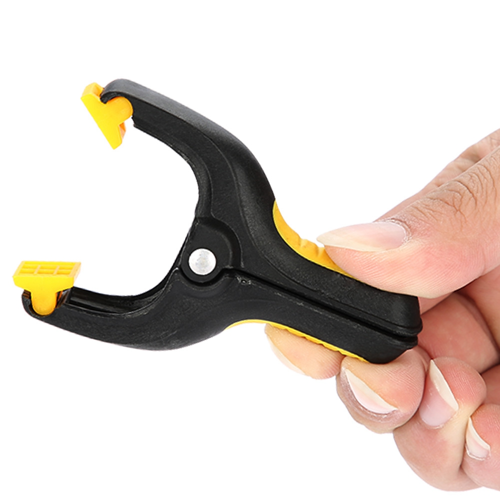 Hand hält eine schwarz-gelbe Plastikklemme. Die Klemme ist offen und bereit, ein Objekt zu greifen.