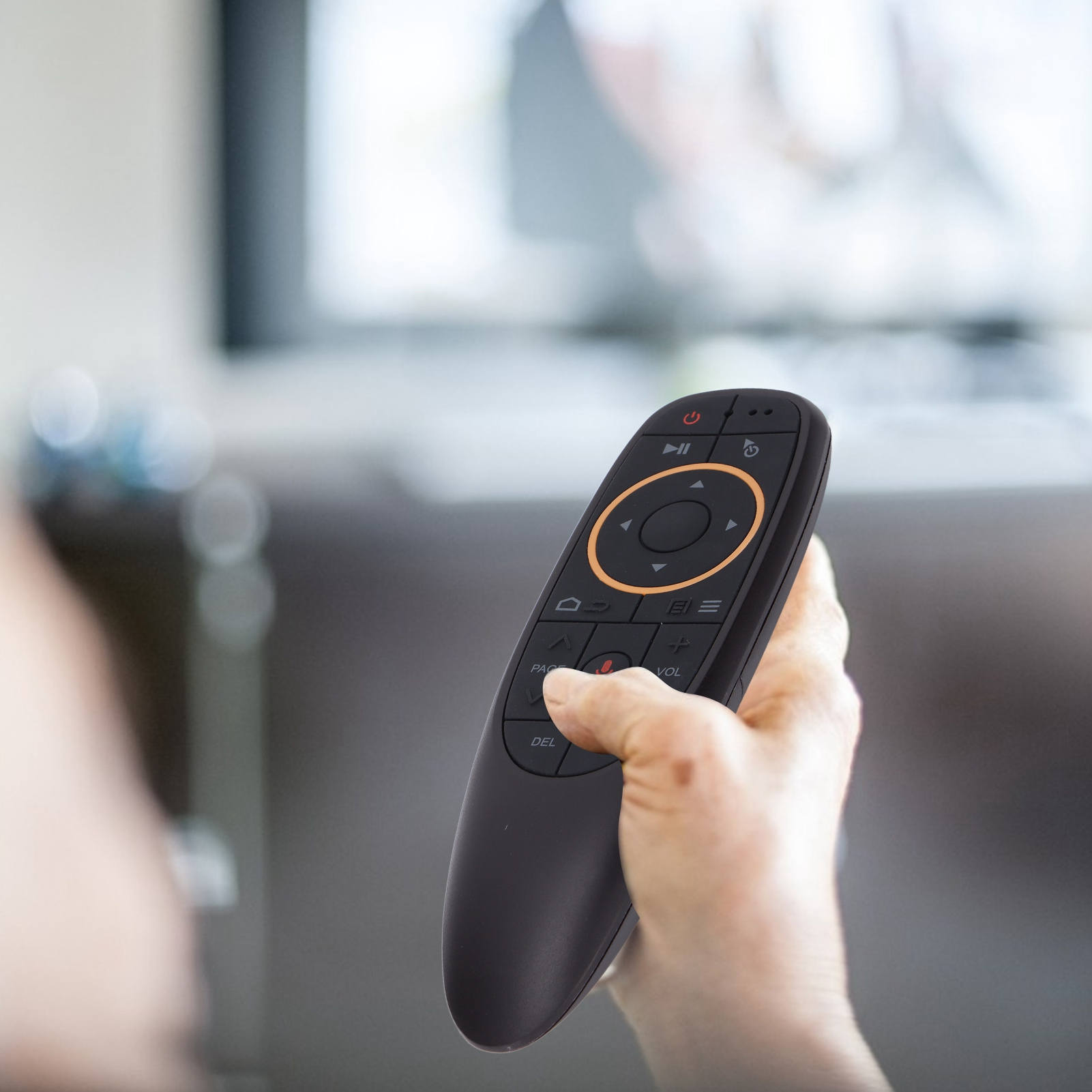 Hand hält eine schwarze Fernbedienung und zeigt sie auf einen Fernseher. Verschwommener Hintergrund zeigt den Fernseher.