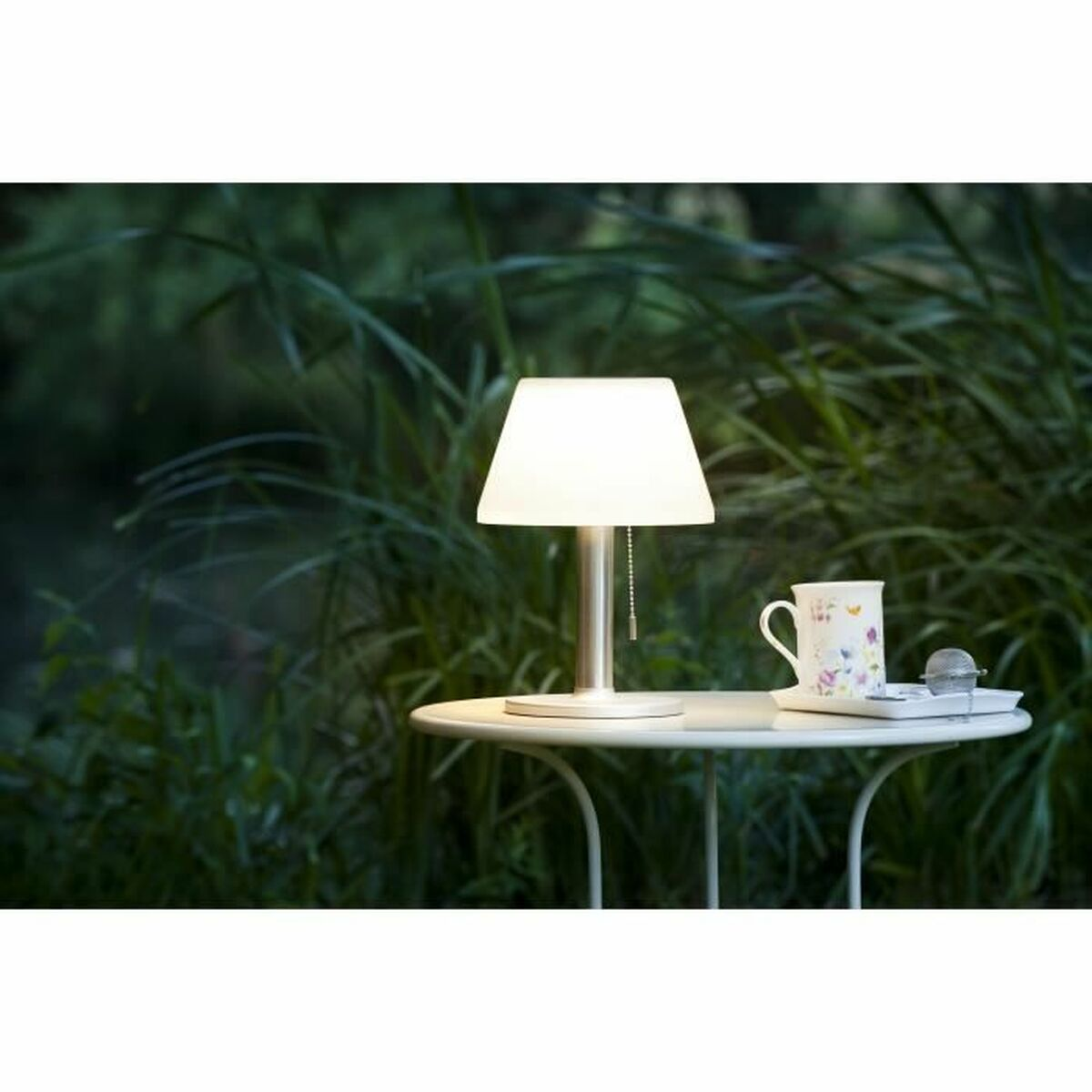 Eine beleuchtete Lampe auf einem Tisch neben einer Tasse und Untertasse, mit grünem Laub im Hintergrund.