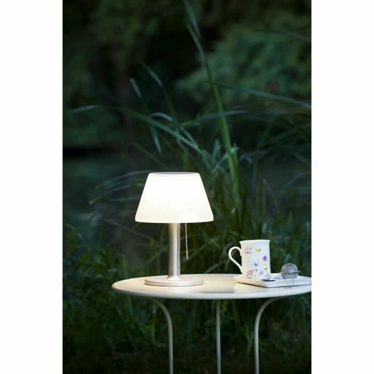 Eine beleuchtete Lampe auf einem Tisch neben einer Tasse und Untertasse, mit grünem Laub im Hintergrund.