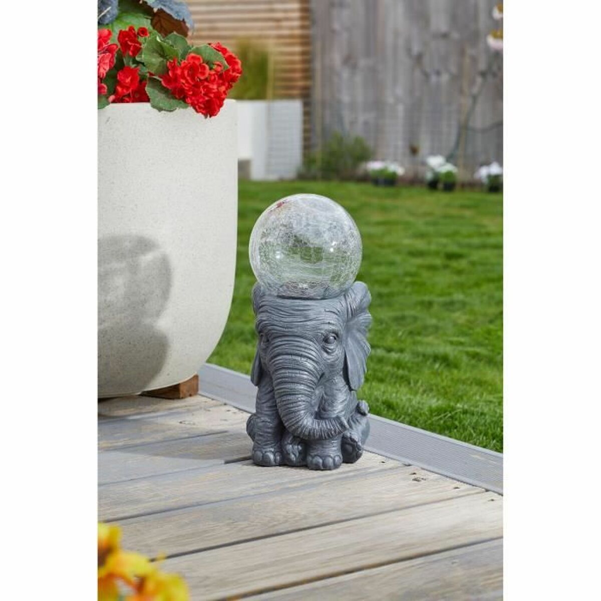 Eine graue Elefantenstatue, die eine klare Glaskugel hält, in einem Garten mit Blumen und Grün.