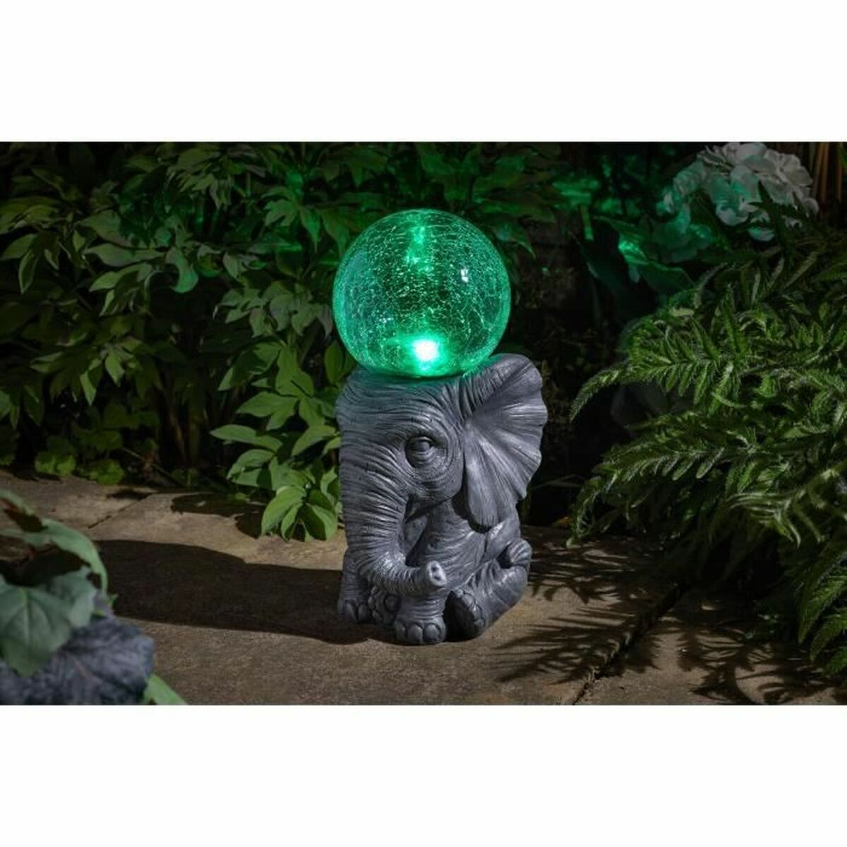 Eine graue Elefantenstatue, die eine leuchtende grüne Glaskugel hält, in einem Garten mit Grün umgeben.