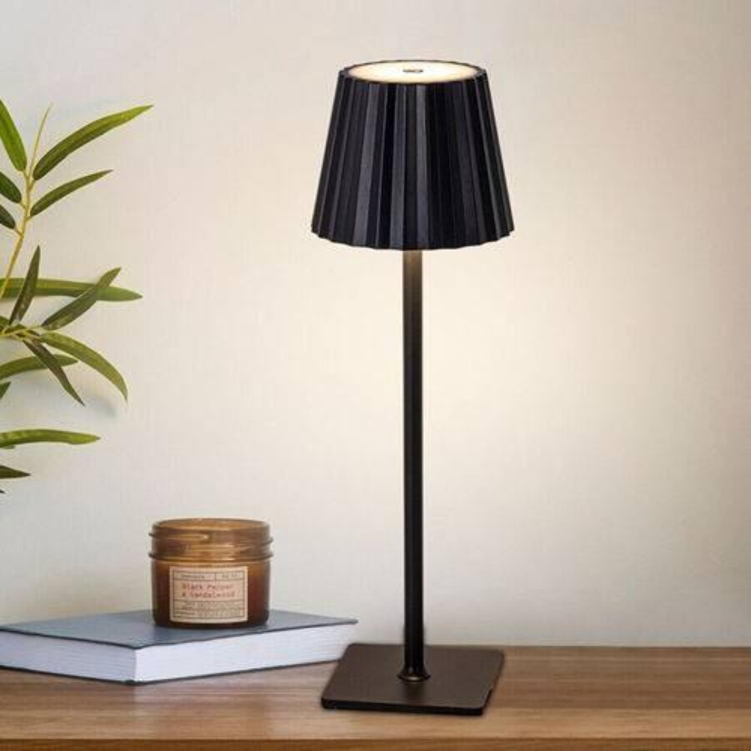 Moderne schwarze Tischlampe mit schwarzem Lampenschirm, auf einem Holztisch mit Buch und Kerze.