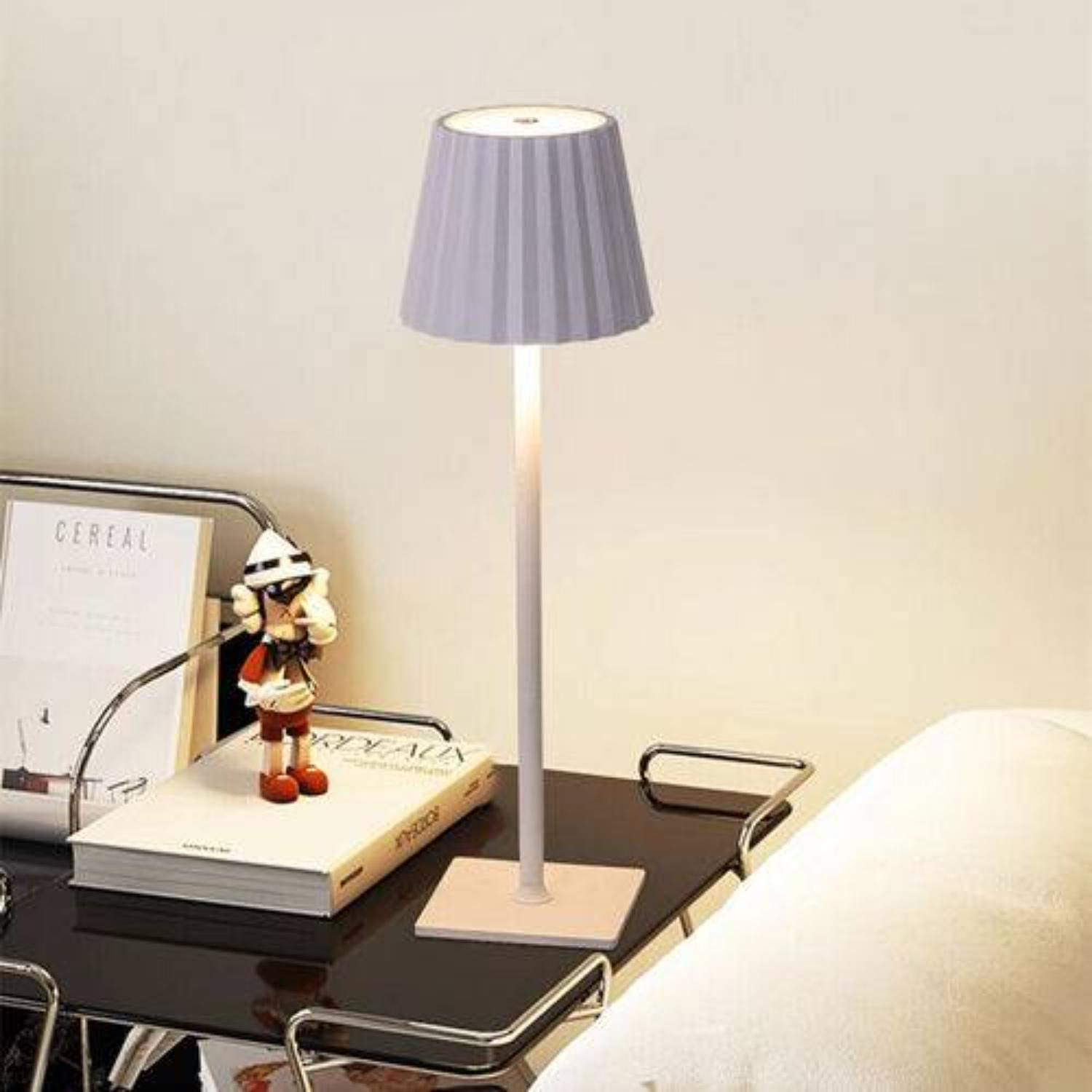Moderne Tischlampe mit hellgrauem Lampenschirm, auf einem Tisch, neben einem Buch und einer dekorativen Figur.