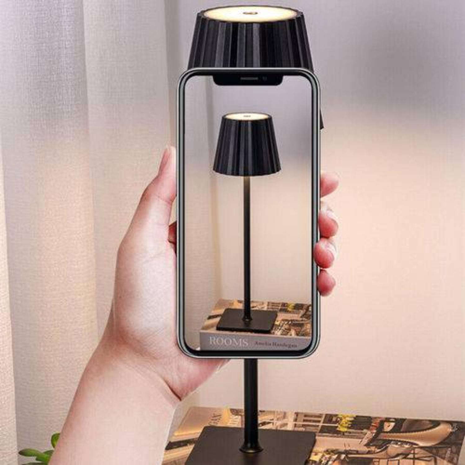 Hand, die ein Telefon hält, das eine Lampe zeigt. Die Lampe ist schwarz mit geripptem Schirm. Das Telefon zeigt die gleiche Lampe.