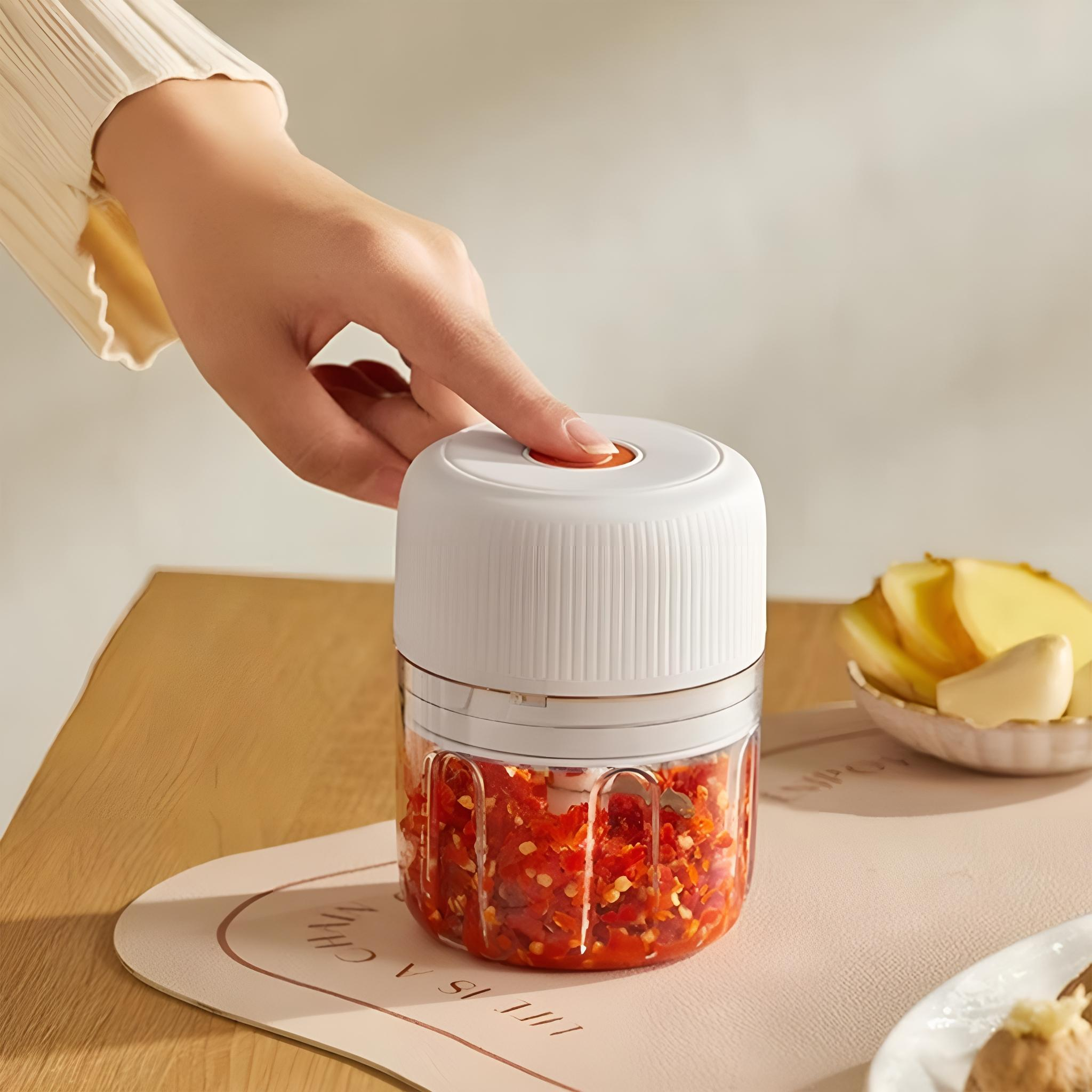 Die Hand einer Person, die einen Knopf an einem kleinen, weißen Zerkleinerer mit gehackten roten Paprika drückt.