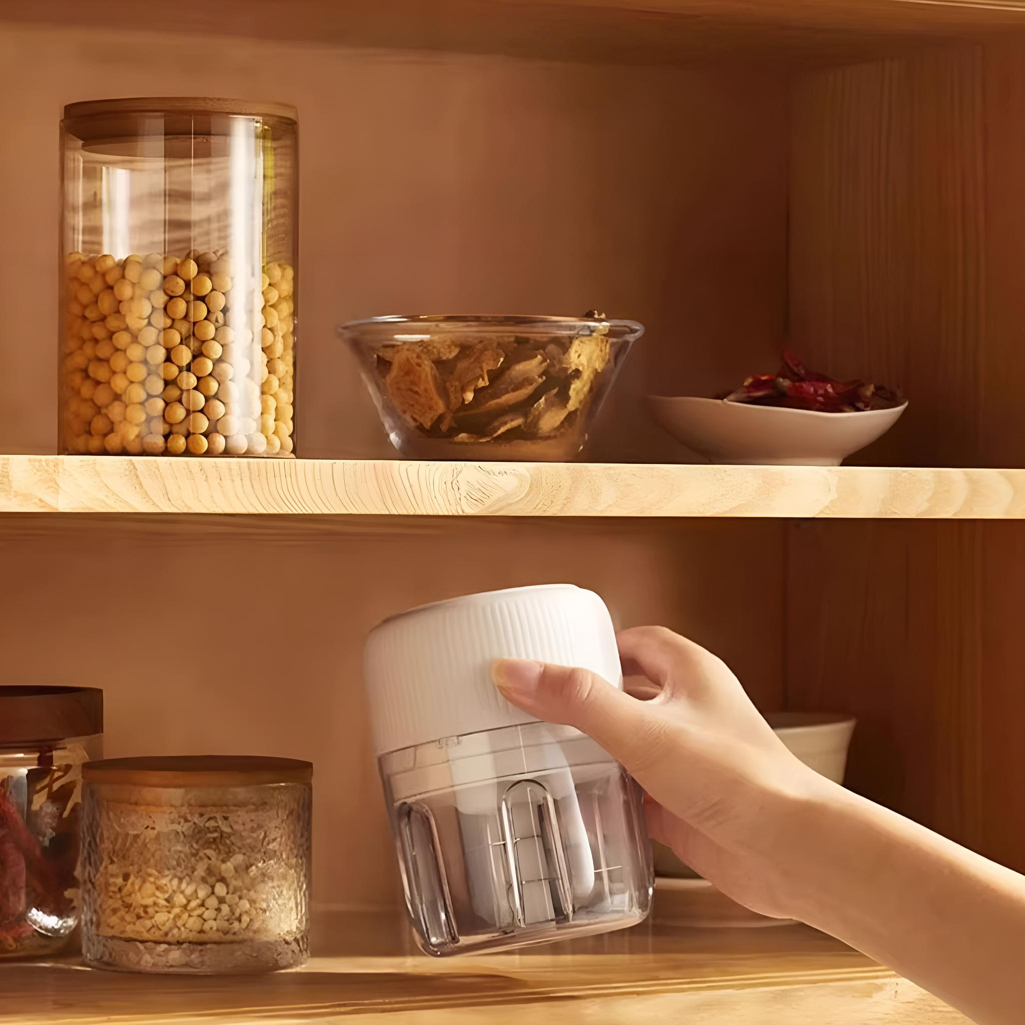 Eine Hand hält einen weißen und transparenten Zerkleinerer in einem Holzschrank. Gläser, Schalen und Zutaten sind sichtbar.