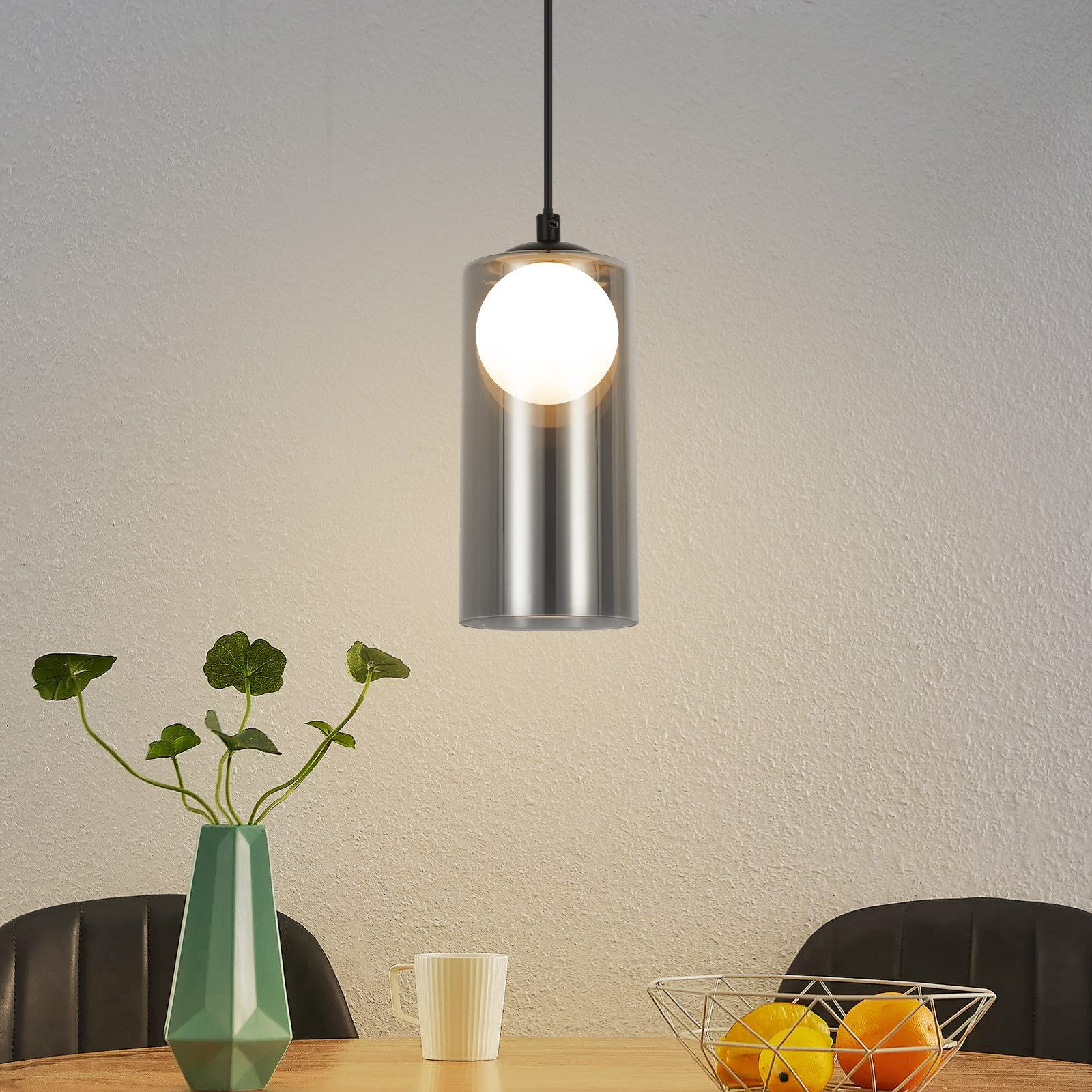 Una lampada a sospensione con un paralume cilindrico in vetro e una lampadina sferica bianca è appesa sopra un tavolo da pranzo.