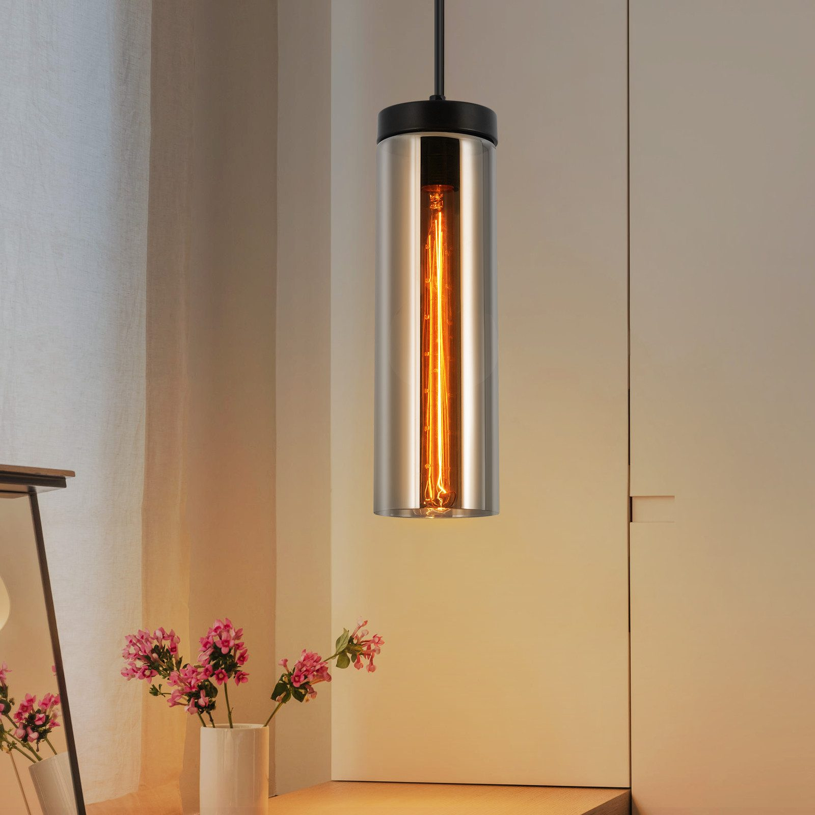 Una singola lampada a sospensione con paralume cilindrico in vetro e lampadina a vista. Fiori rosa nel vaso presenti.