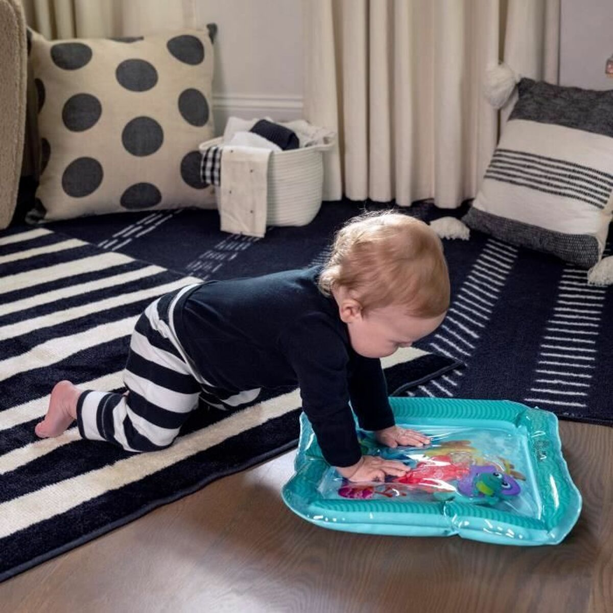 Baby spielt mit Wassermatte auf blau gestreiftem Teppich, Kissen im Hintergrund.