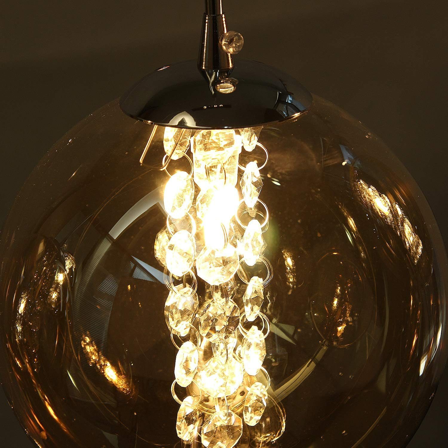 Primo piano di una lampada a sospensione sferica in vetro ambra con dettagli di cristallo, illuminata dall'interno, appesa a una base cromata.