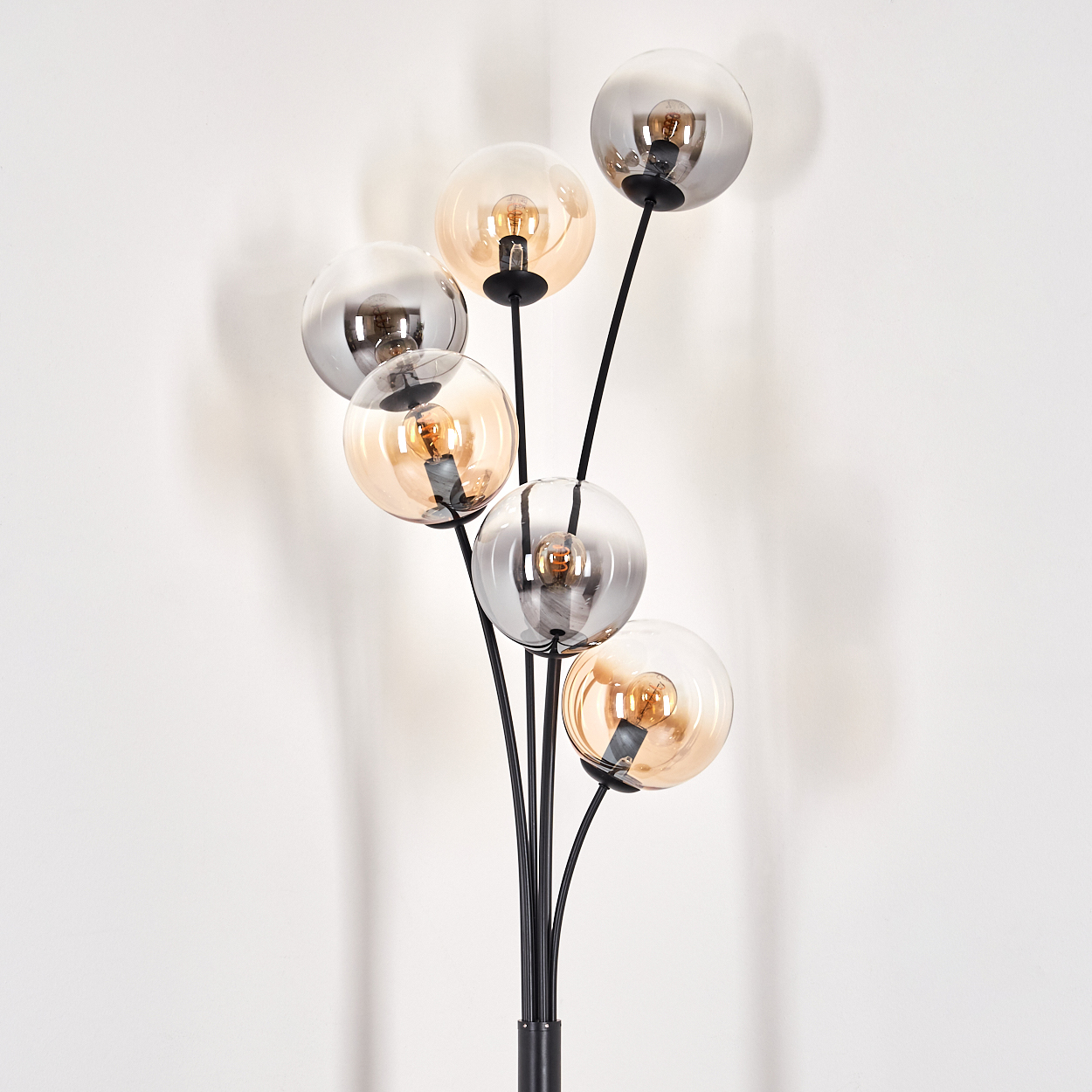 Eine schwarze Stehlampe mit sechs Glaskugeln. Die Lampe ist eingeschaltet und steht vor einer weißen Wand.
