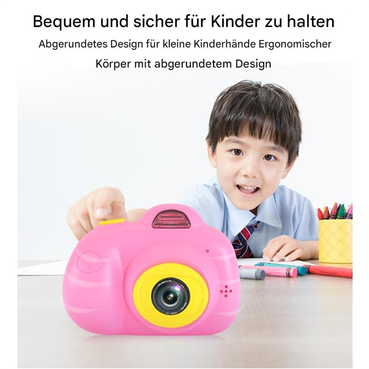 Ein junger Junge lächelt und hält eine rosa Kinderkamera mit gelbem Objektiv vor schlichtem Hintergrund.
