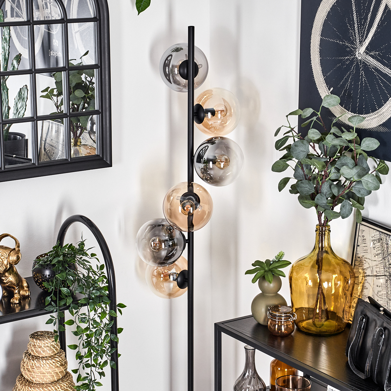 Schwarze Stehlampe mit bernsteinfarbenen und grauen Glaskugeln. Hintergrund zeigt Raumdekor, Pflanzen und einen schwarzen Bogen.