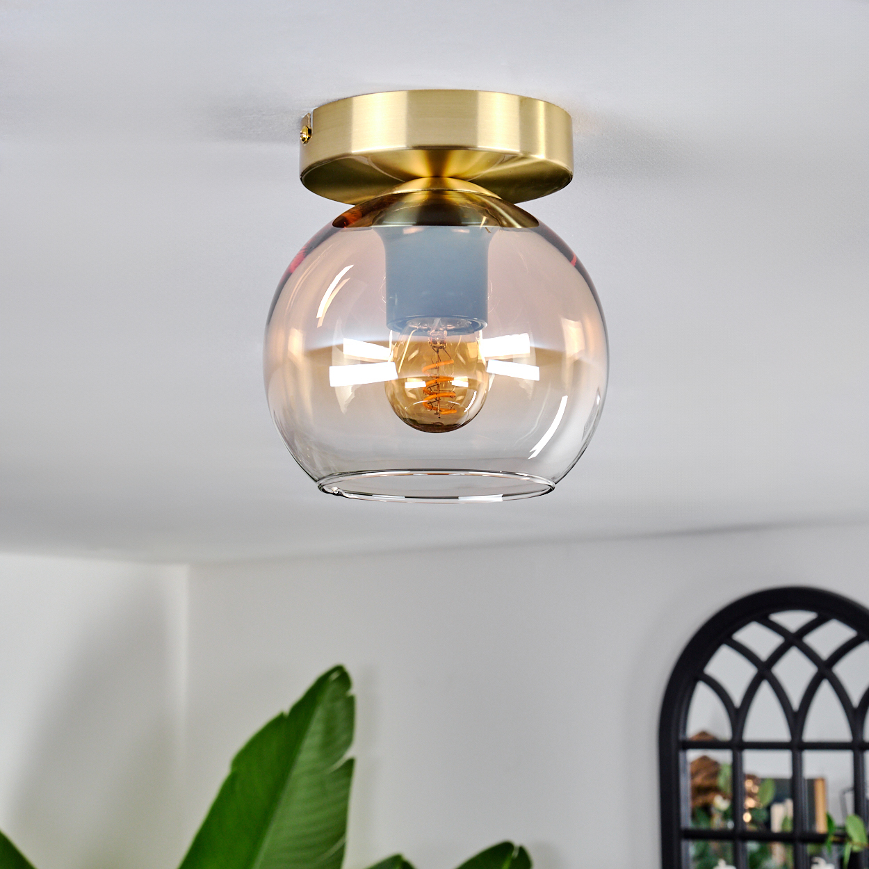 Deckenleuchte mit goldfarbenem Finish und bernsteinfarbenem Glasschirm, Glühbirne sichtbar. Weiße Decke und Raumdetails im Hintergrund.