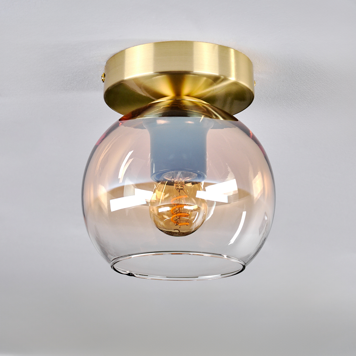 Eine Deckenlampe mit goldener Basis und Glaskugel. Die Glühbirne ist sichtbar. Weißer Hintergrund.