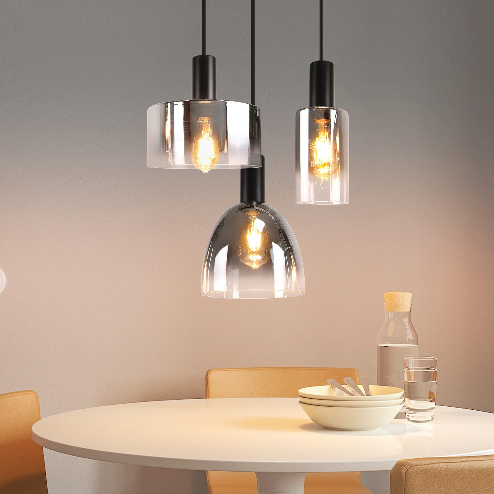 Tre lampade a sospensione con forme diverse, paralumi in vetro ambrato e lampadine accese, appese sopra un tavolo da pranzo.