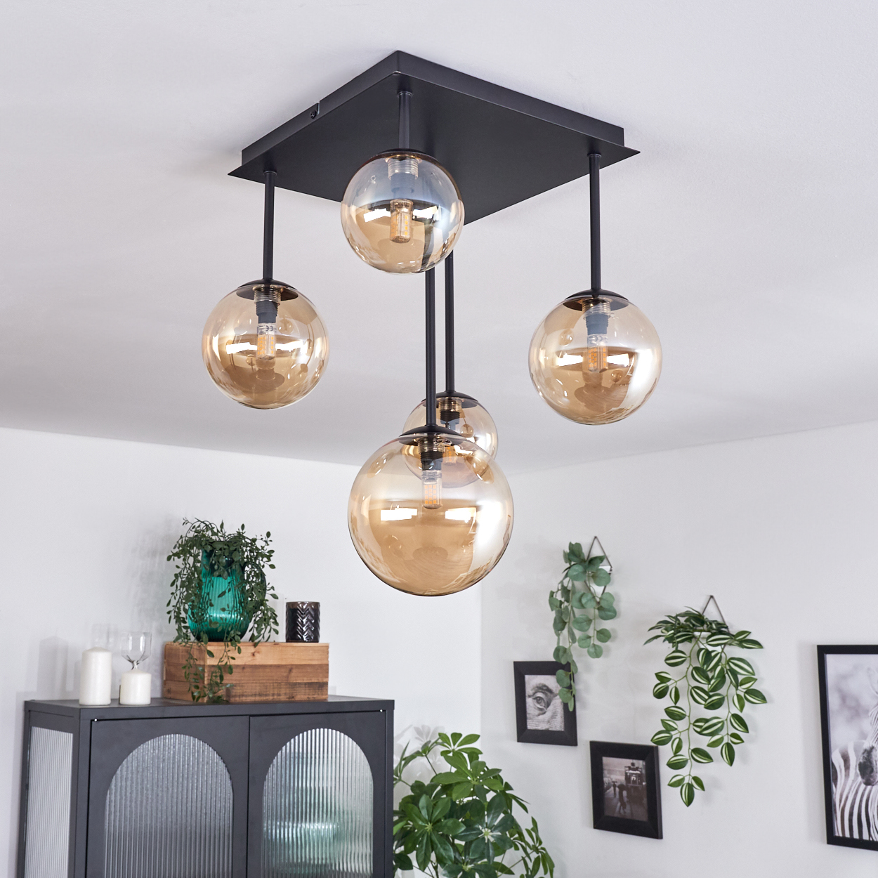 Deckenlampe mit 4 bernsteinfarbenen Kugeln. Quadratischer schwarzer Sockel und schwarze Stangen. Weiße Decke und Wände.