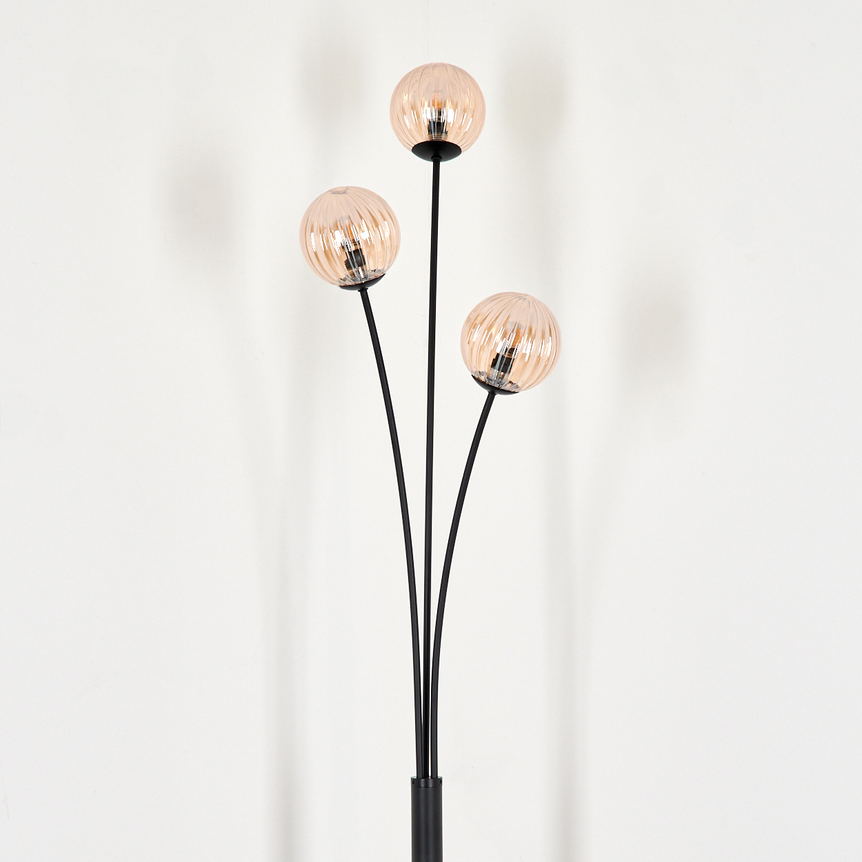 Eine Stehlampe mit drei rosa Kugelschirmen auf schwarzen Stielen vor einer weißen Wand.