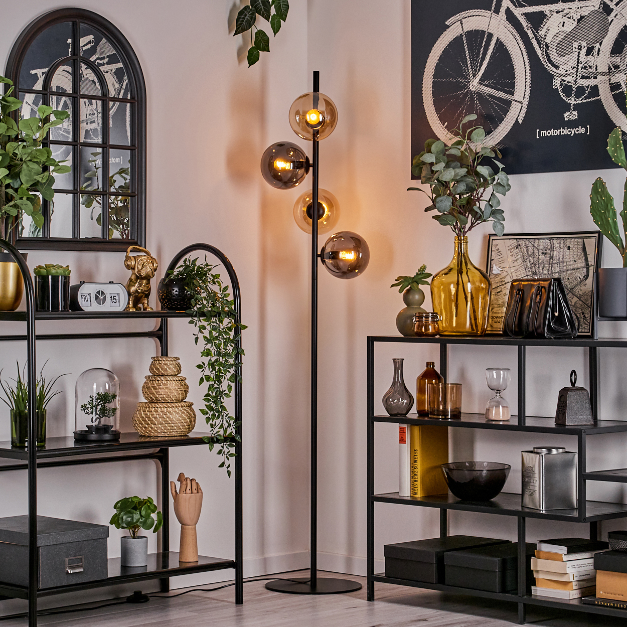 Eine schwarze Stehlampe mit vier Glaskugeln, die in einem Wohnzimmer mit Regalen, Pflanzen und Dekorationsartikeln steht.