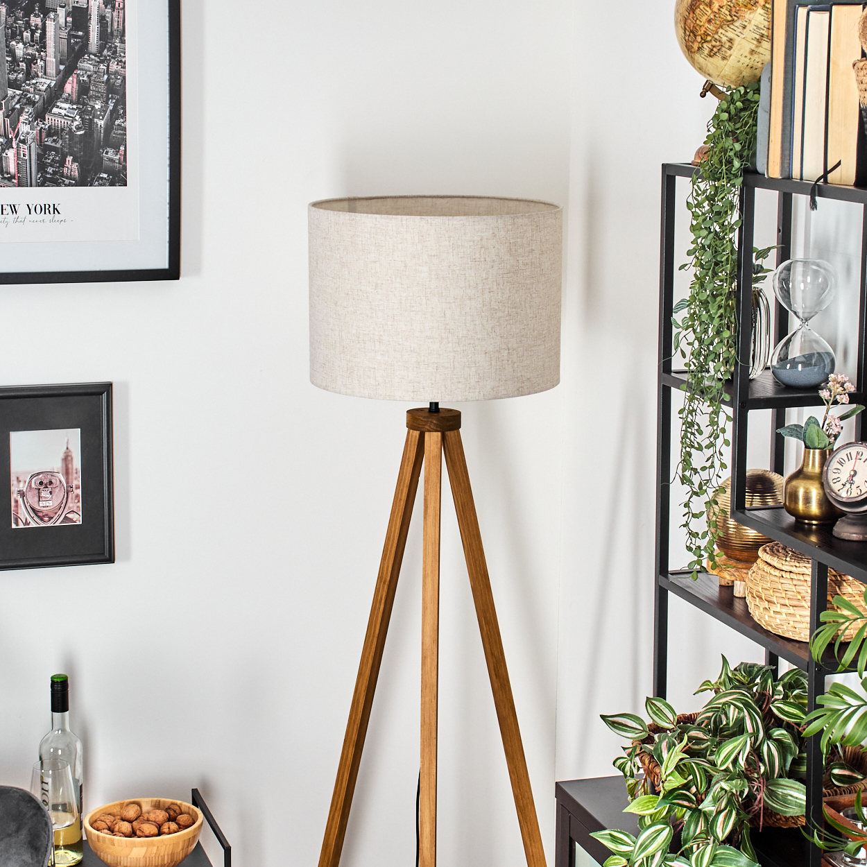 Eine hölzerne Stativ-Stehlampe mit einem beigefarbenen zylindrischen Lampenschirm, in einem Raum mit Regal und Dekoration.