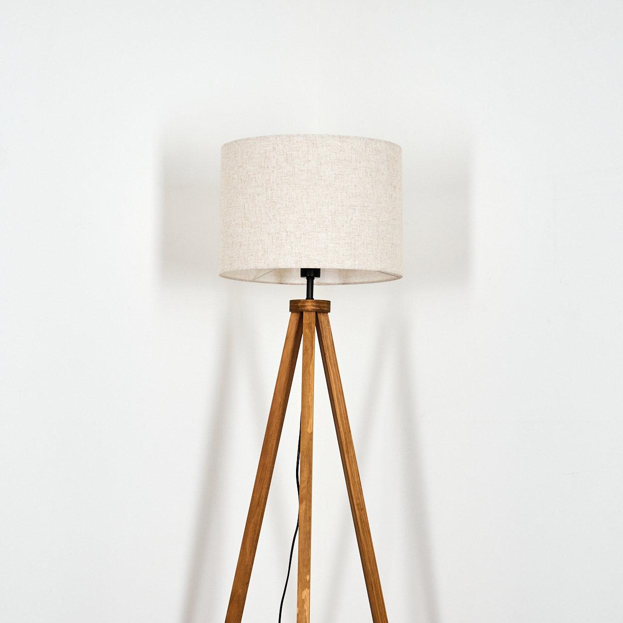 Holz-Stativ-Stehlampe mit beigem zylindrischen Schirm. Der Hintergrund ist eine schlichte weiße Wand.