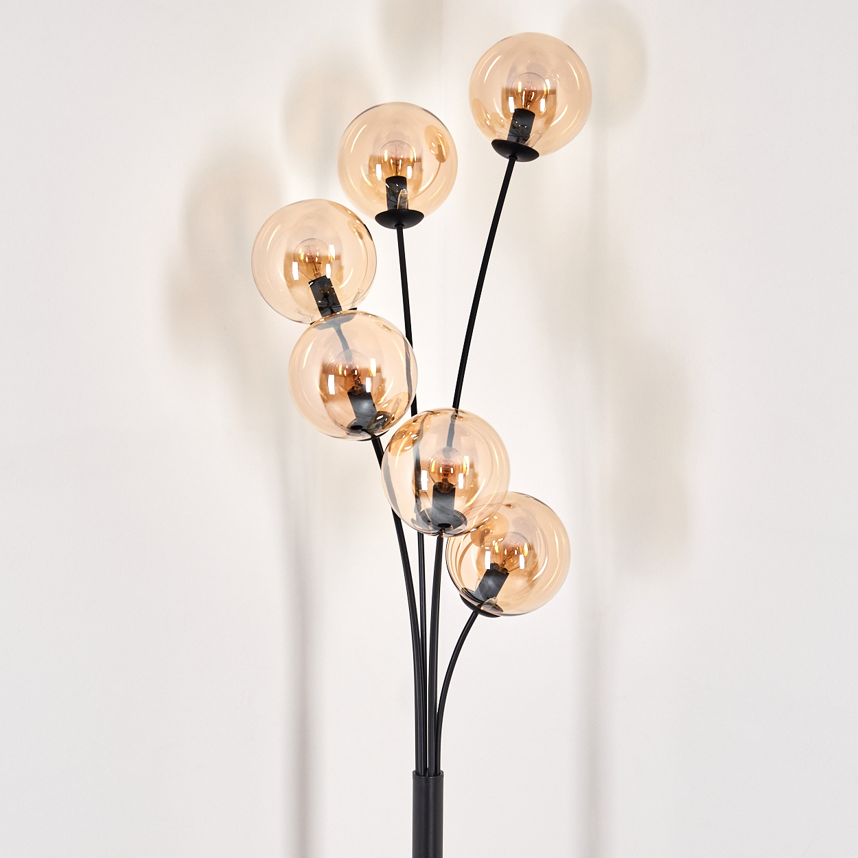 Stehlampe mit sechs bernsteinfarbenen Glaskugeln. Schwarzer Stiel und Sockel. Vor einer weißen Wand.