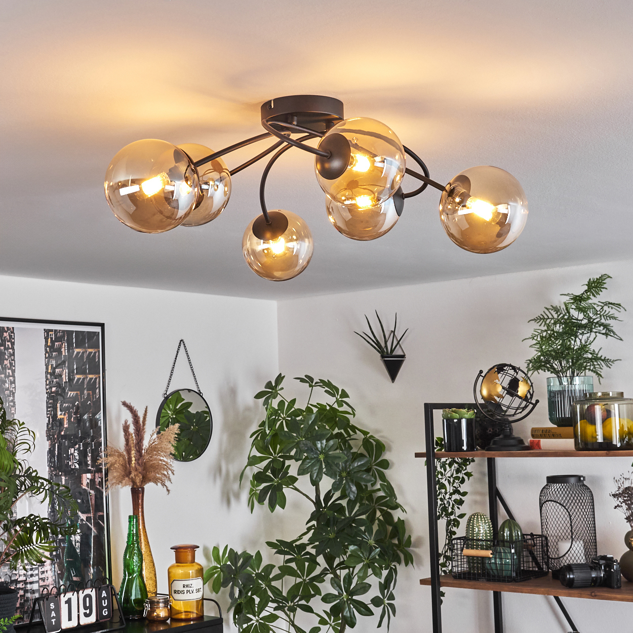 Deckenlampe mit bernsteinfarbenen Glaskugeln, schwarzem Rahmen, Pflanzen und Möbeln im Hintergrund.