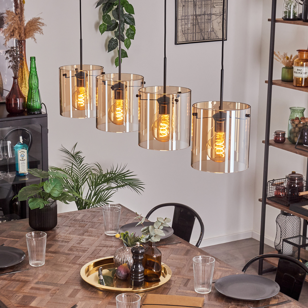 Esszimmer mit vier bernsteinfarbenen Pendelleuchten, Holztisch, Tellern, Gläsern und Pflanzen im Hintergrund.