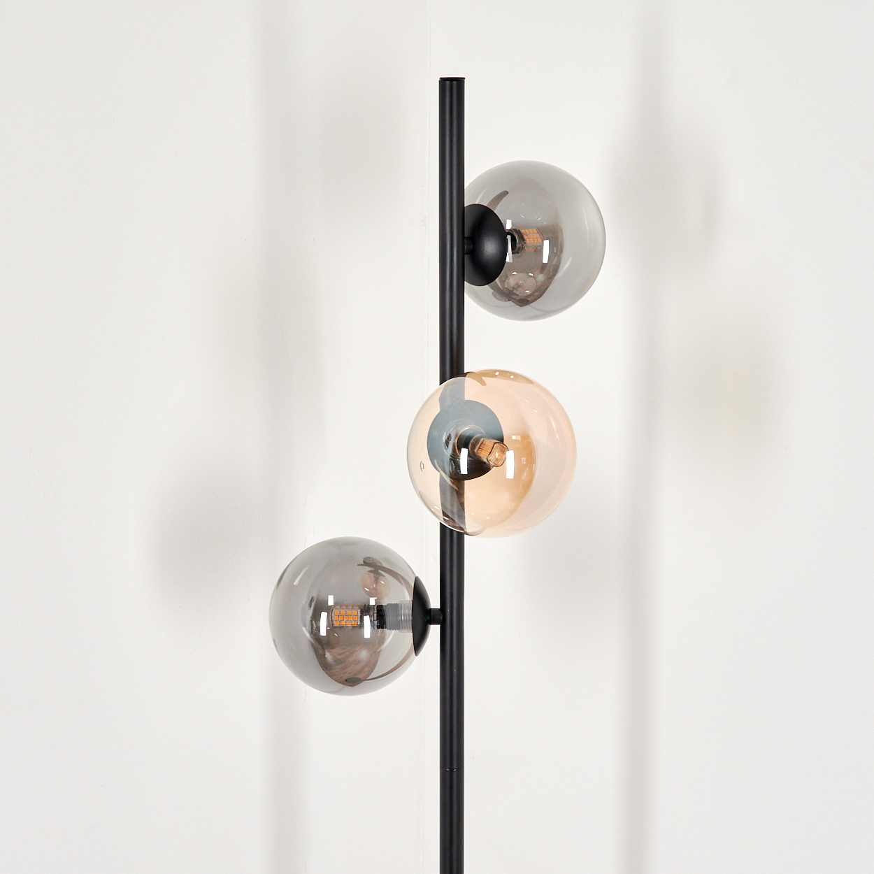 Eine schwarze Stehlampe mit drei Glaskugeln, zwei grauen und einer bernsteinfarbenen. Die Lampe steht an einer weißen Wand.