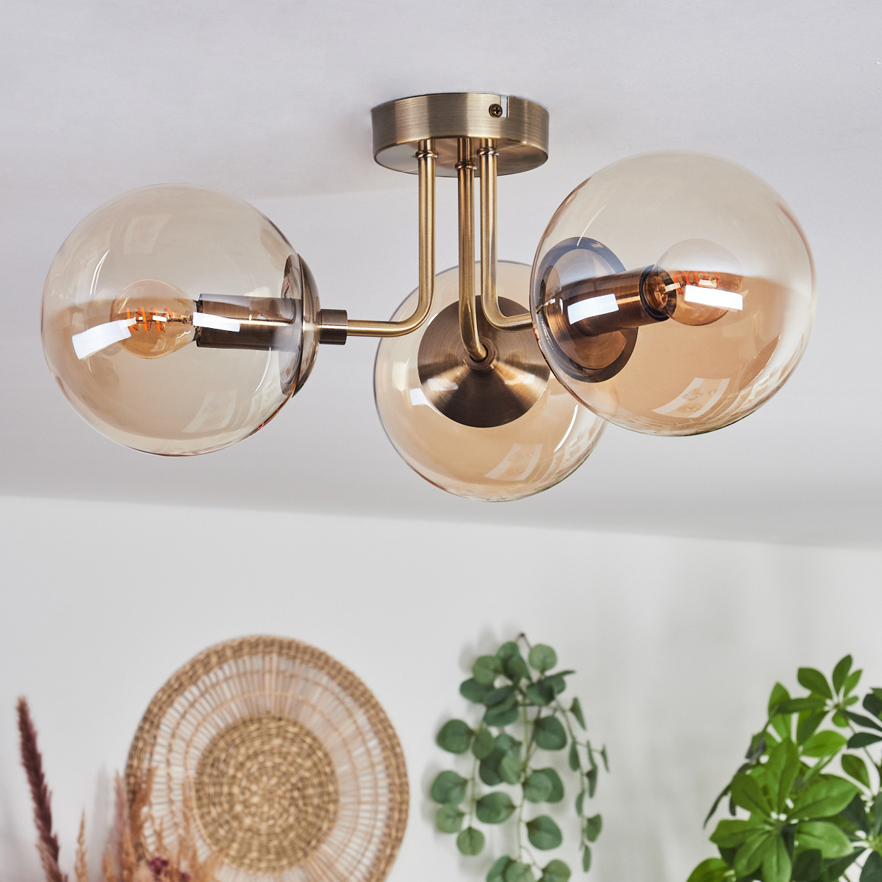 Eine Deckenlampe mit drei bernsteinfarbenen Glaskugeln. Ein gewebter Teller, eine Pflanze und eine weiße Decke sind zu sehen.