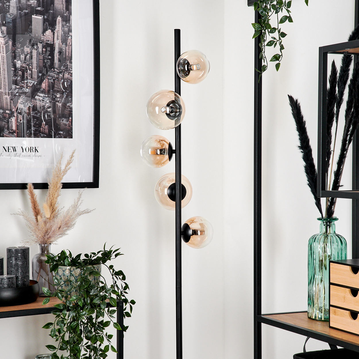 Interieur mit einer schwarzen Stehlampe mit bernsteinfarbenen Kugeln. Enthält Pflanzen, Regale und ein New York-Poster.