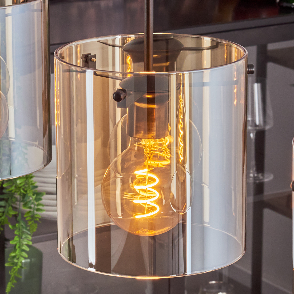 Nahaufnahme einer Pendelleuchte mit bernsteinfarbenem Glaszylinder und glühender Wendelglühbirne im Inneren.