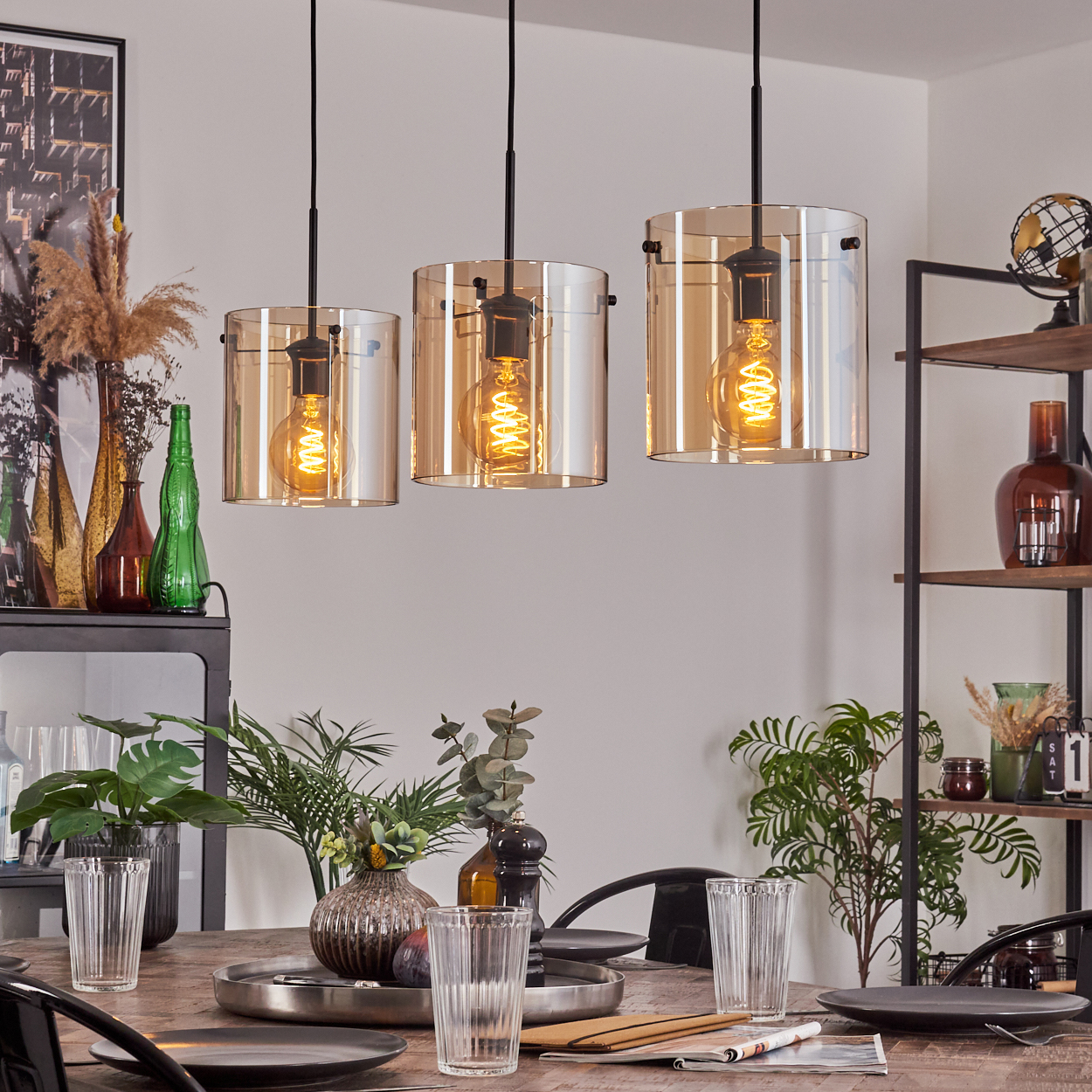 Esszimmeransicht mit drei Pendelleuchten mit bernsteinfarbenen Glasschirmen über einem Holztisch mit Gläsern.