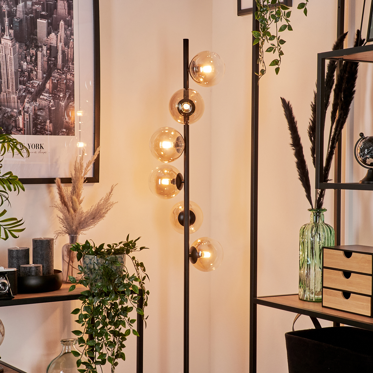 Eine hohe schwarze Lampe mit Kugellampen, umgeben von Dekoration: Regale, Pflanzen und ein Stadtposter.