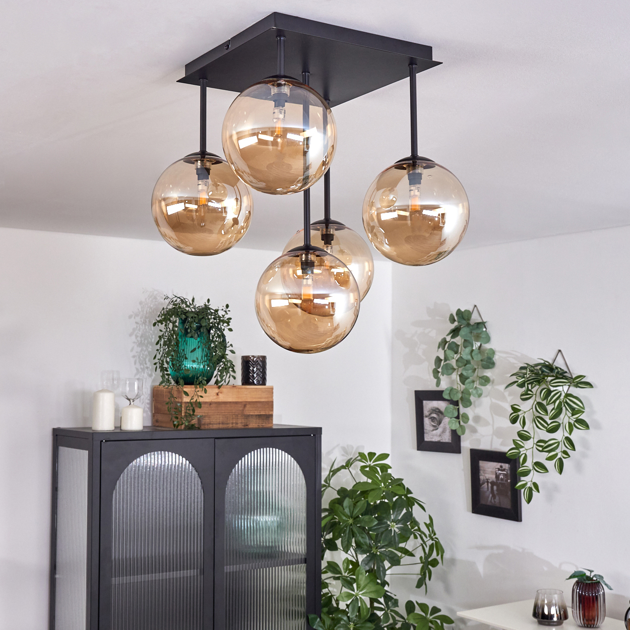 Deckenlampe mit fünf bernsteinfarbenen Glaskugeln. Schwarzer Rahmen. Schrank und Pflanzen im Hintergrund.