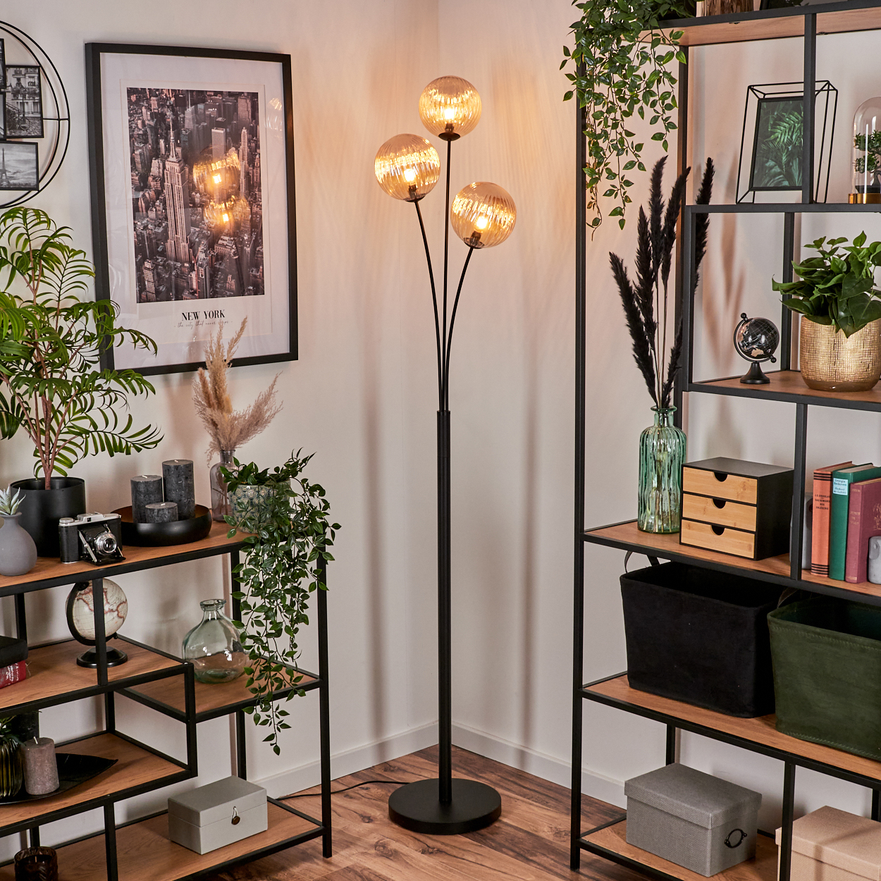 Ein Raum mit einer schwarzen Stehlampe mit drei orangefarbenen Glaskugeln. Regale mit Deko sind vorhanden.