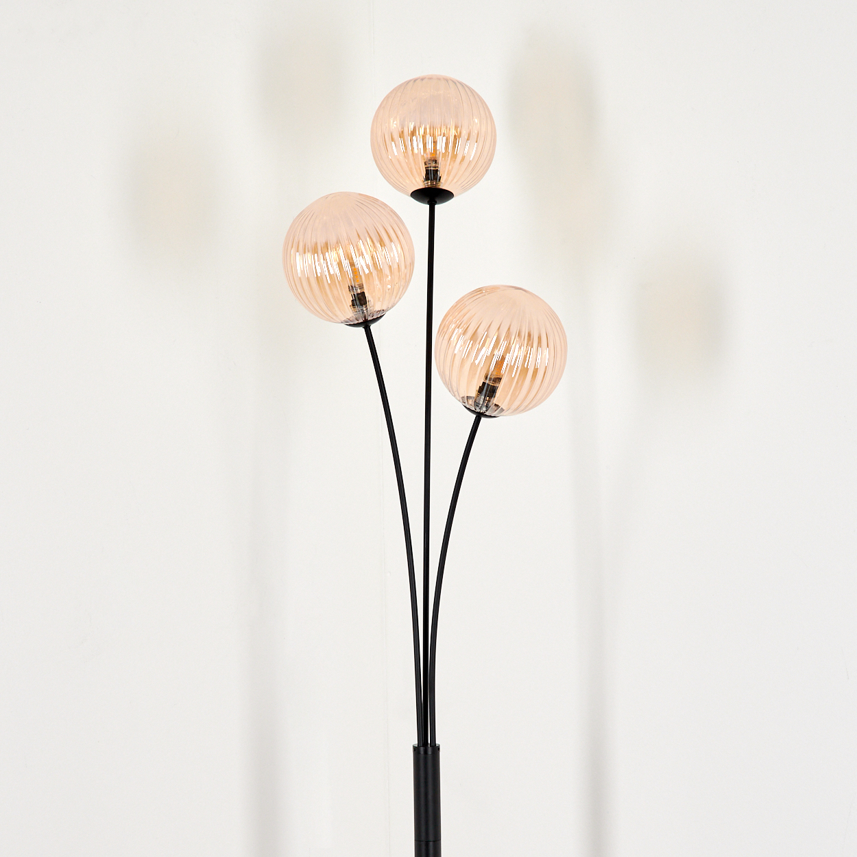 Eine schwarze Stehlampe mit drei orangefarbenen Glaskugeln. Der Hintergrund ist eine weiße Wand.