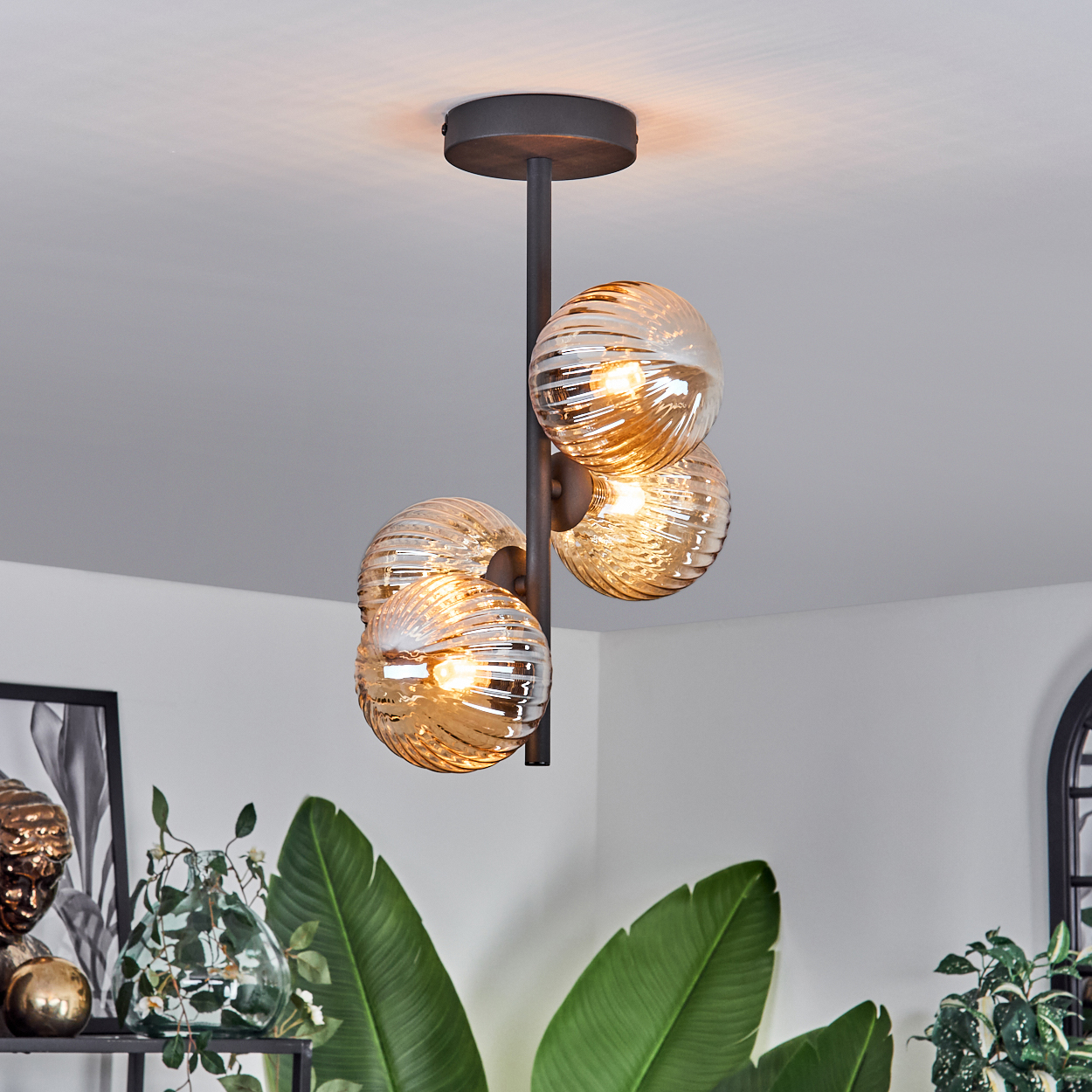 Eine schwarze Deckenlampe mit drei geriffelten bernsteinfarbenen Glaskugelschirmen, vor weißem Hintergrund.
