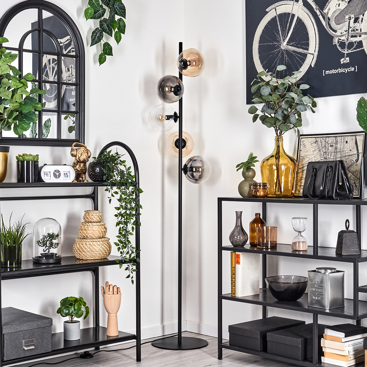 Eine Zimmerecke mit einer schwarzen Stehlampe, Regalen und Dekoration. Die Lampe hat Kugelschirme, die Regale halten Pflanzen und Objekte.