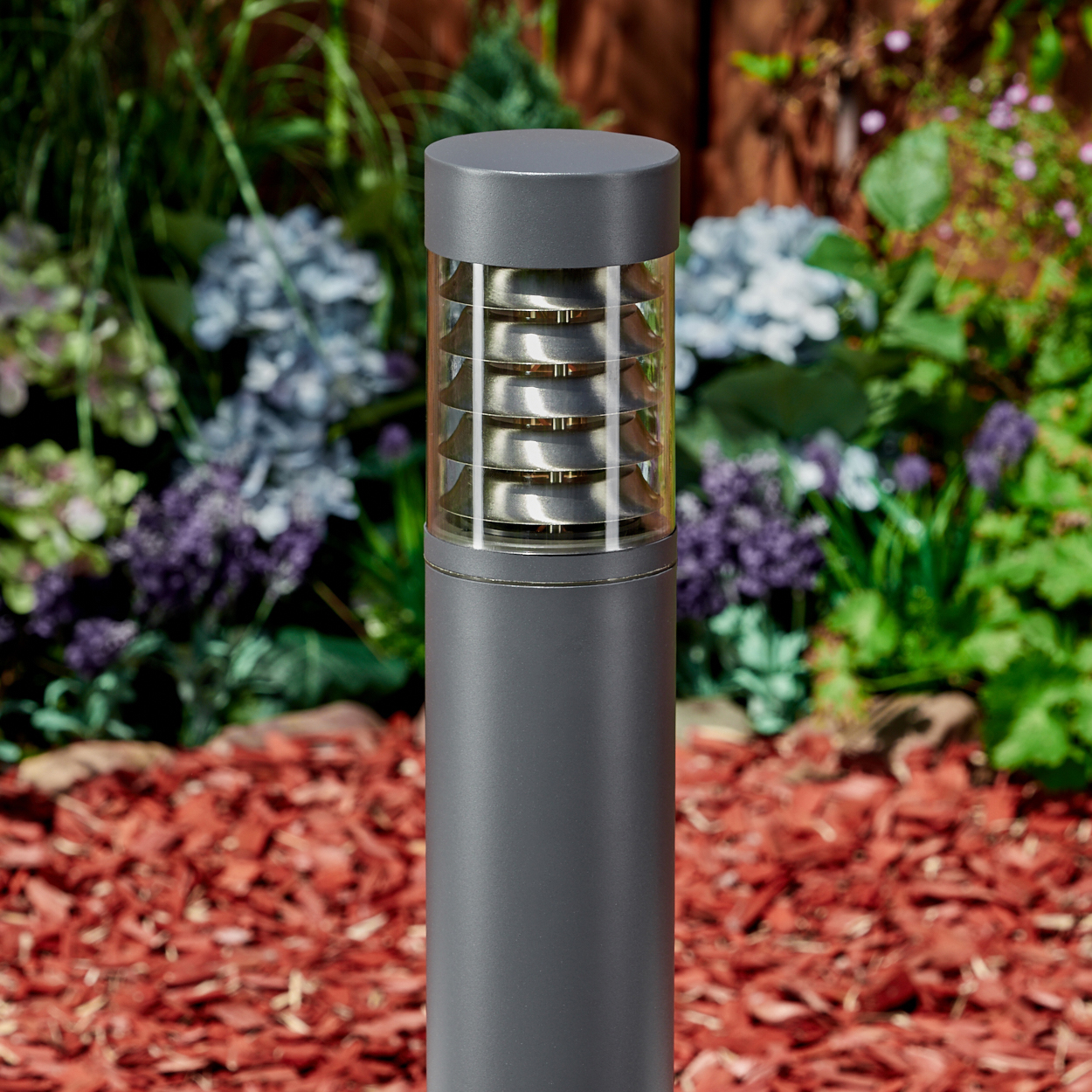 Eine dunkelgraue, zylindrische Außenlampe mit einem Glasabschnitt und Gartenhintergrund.