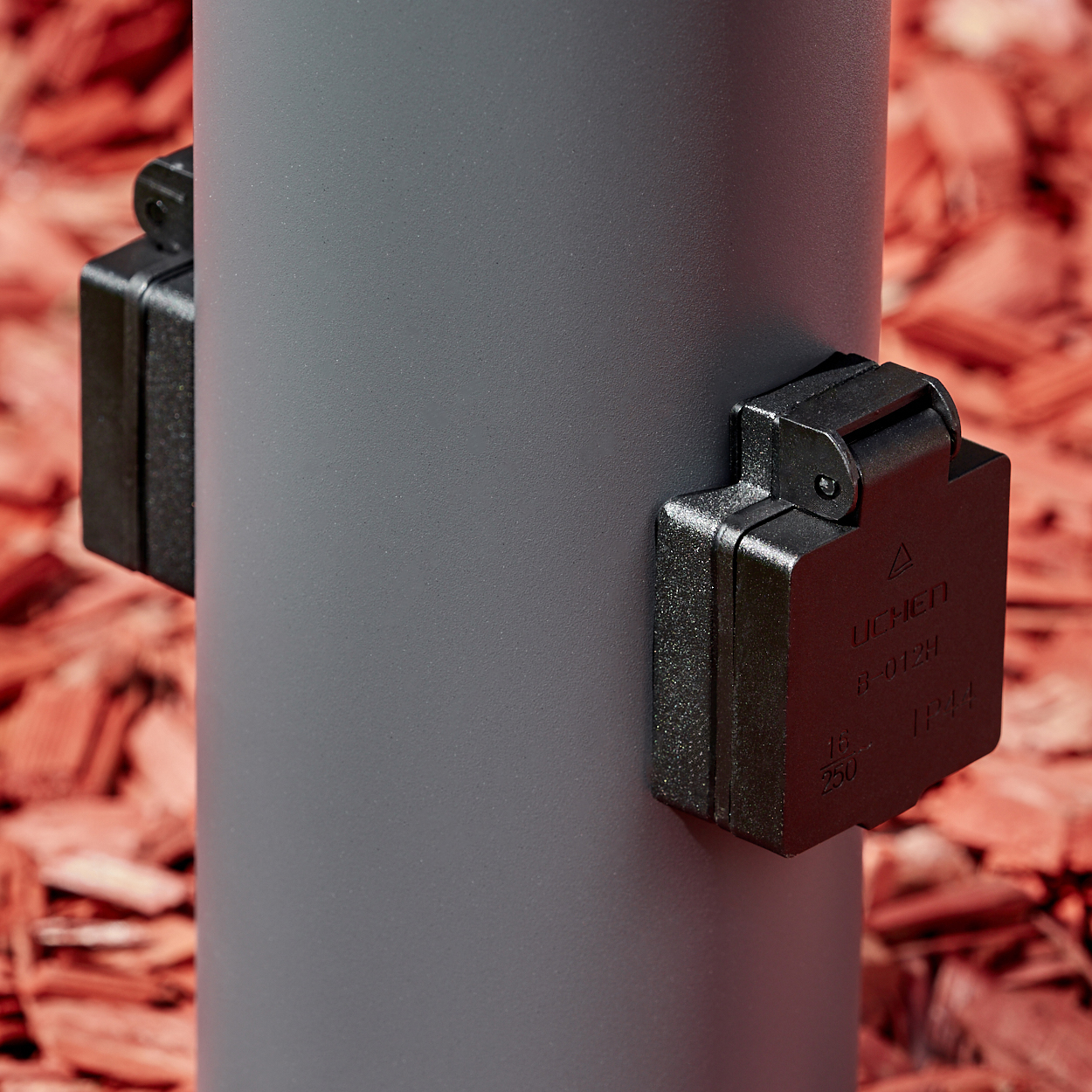 Nahaufnahme der beiden schwarzen Steckdosen einer grauen Außenlampe. Roter Mulch-Hintergrund.