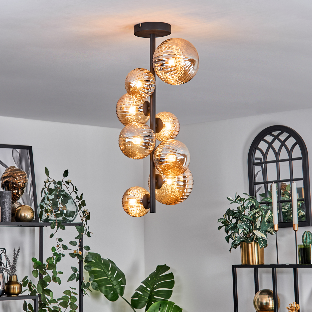 Deckenlampe mit mehreren bernsteinfarbenen Glaskugeln. Die Lampe befindet sich in einer Raumeinstellung in der Nähe von Pflanzen und Dekorationen.