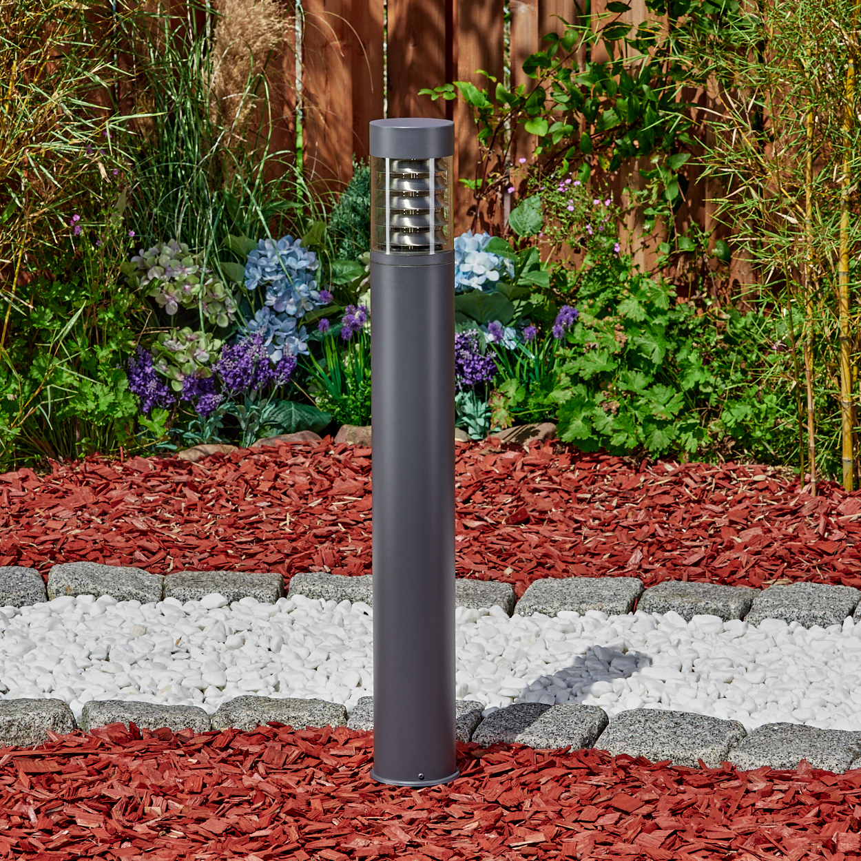 Außenleuchte in einem Garten, mit roter Mulch, weißen Steinen und Pflanzen im Hintergrund.