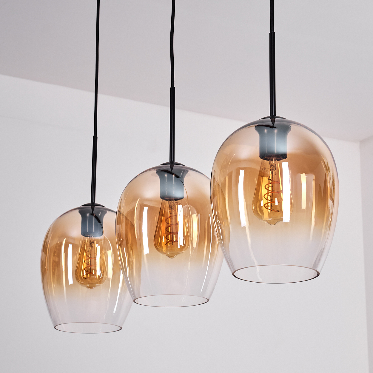 Drei Pendelleuchten mit bernsteinfarbenen Glasschirmen, schwarzen Kabeln und sichtbaren Glühbirnen vor weißer Decke.