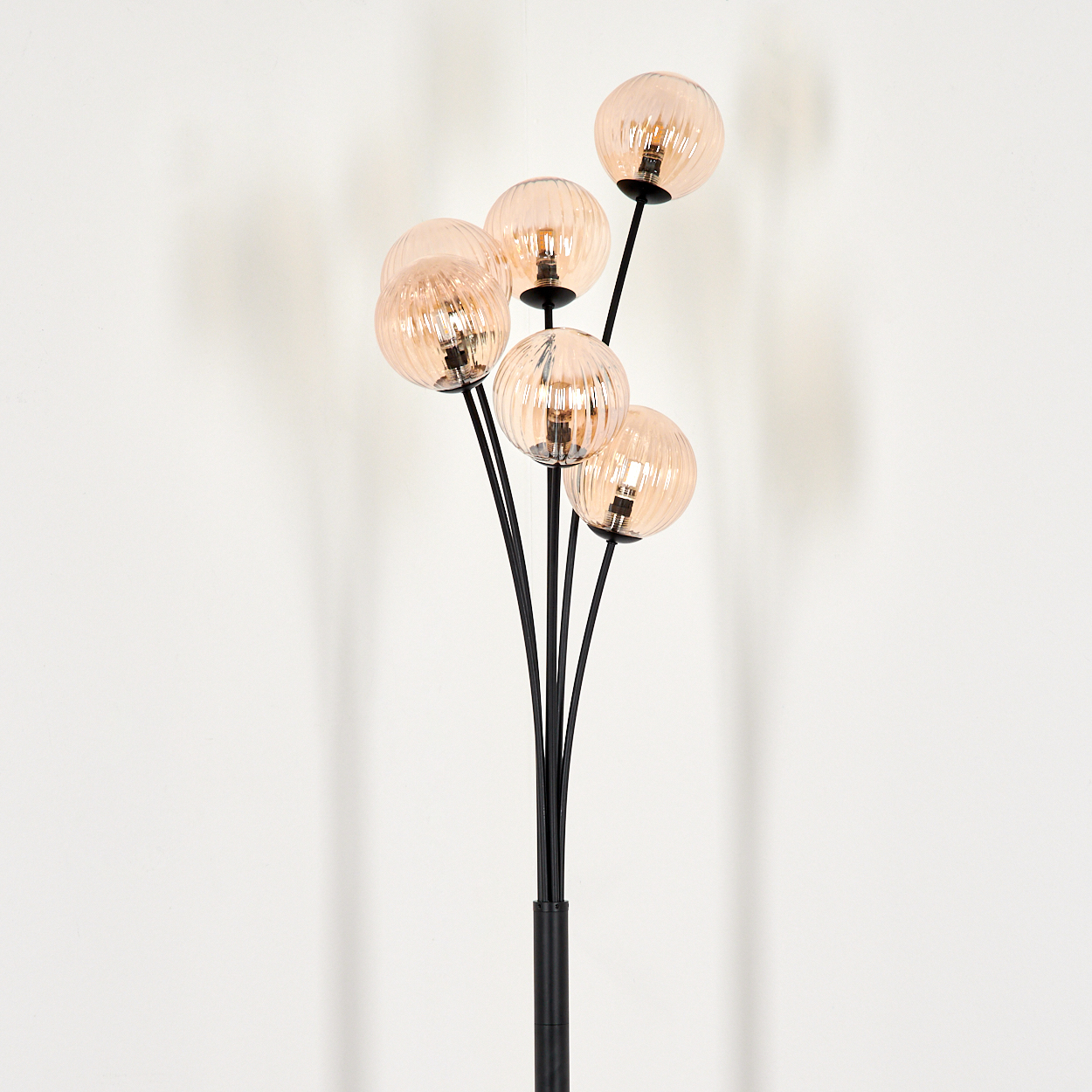 Eine schwarze Stehlampe mit sechs bernsteinfarbenen Glaskugelschirmen an gebogenen Stielen vor einer weißen Wand.